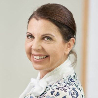 Frau mit dunklem langem Haar, das zu einem Zopf zusammengebunden ist, lächelt in die Kamera.