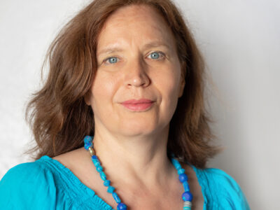 Frau mit langem, hellbraunem Haar. Sie trägt eine blaue Bluse und blaue Halskette.