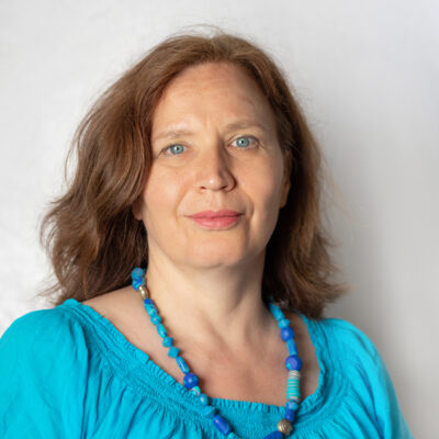 Frau mit langem, hellbraunem Haar. Sie trägt eine blaue Bluse und blaue Halskette.