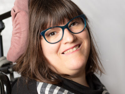 Lächelnde Frau mit Brille. Sie hat längeres, braunes Haar. Im Hintergrund eine Kopfstütze.