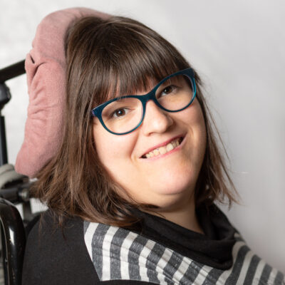 Lächelnde Frau mit Brille. Sie hat längeres, braunes Haar. Im Hintergrund eine Kopfstütze.