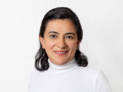 Frau mit schulterlangem dunkelbraunen Haar und weißem Rollkragen-Oberteil lächelt in die Kamera