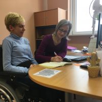Eine blonde kurzhaarige Frau im Rollstuhl sitzt mit einer langhaarigen Fraun mit Brille an einem Schreibtisch. Die Frau mit Brille schreibt in ein Buch.