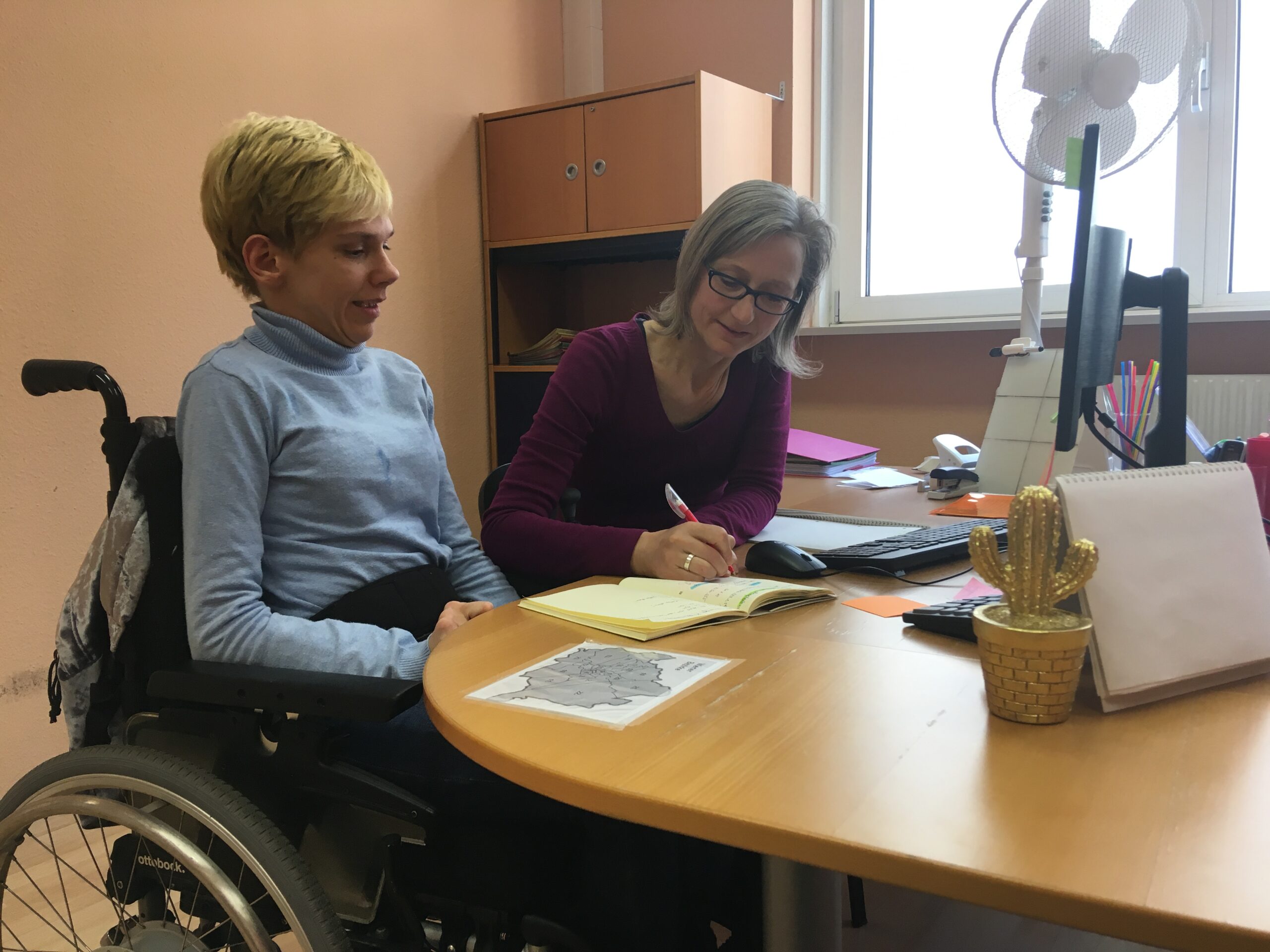 Eine blonde kurzhaarige Frau im Rollstuhl sitzt mit einer langhaarigen Fraun mit Brille an einem Schreibtisch. Die Frau mit Brille schreibt in ein Buch.