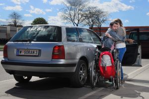 Ein Rollstuhl steht neben einem Auto. Ein Mann stützt sich an einer Person ab. Beide stehen neben der Autotür.