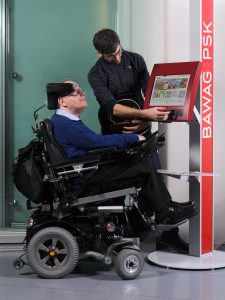 Ein Mann im Rollstuhl steht vor einem Bankomaten. Ein zweiter Mann bedient diesen.