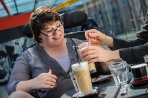 Lachende Frau vor 2 Kaffee-Gläsern. Zwei Hände halten ihr das Glas mit Strohhalm hin.