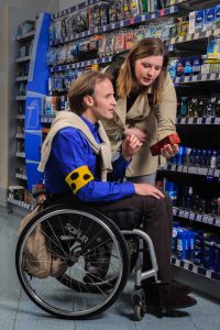 Drogerie: Eine Frau hält einem Mann im Rollstuhl mit Blindenschleifeein Parfum hin. Sie scheint ihm etwas zu erklären.