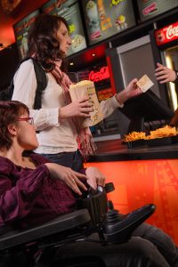 Kino: Frau im Rollstuhl steht beim Popcorn. Stehende Frau hält Popcorn und Gelschein (zum Bezahlen) in der Hand.