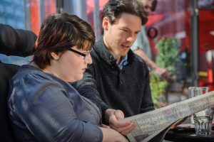 Zwei Personen mit einer Zeitung. Ein junger Mann hält diese. beide scheinen zu lesen.