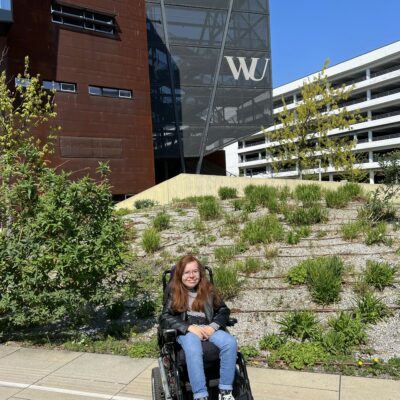 Frau im Rollstuhl. Dahinter die Gebäude der Wirtschaftsuniversität Wien.
