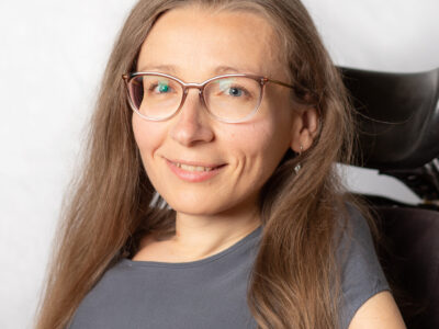 Frau mit langem, blondem Haar und Brille. Mit Lehne des Rollstuhls im Hintergrund.