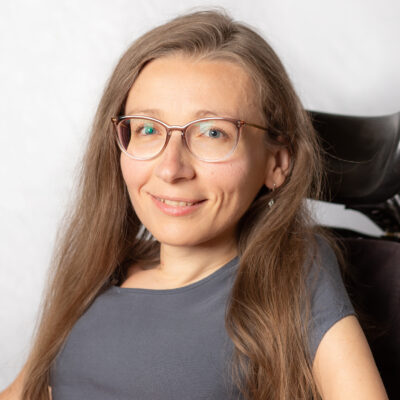 Frau mit langem, blondem Haar und Brille. Mit Lehne des Rollstuhls im Hintergrund.