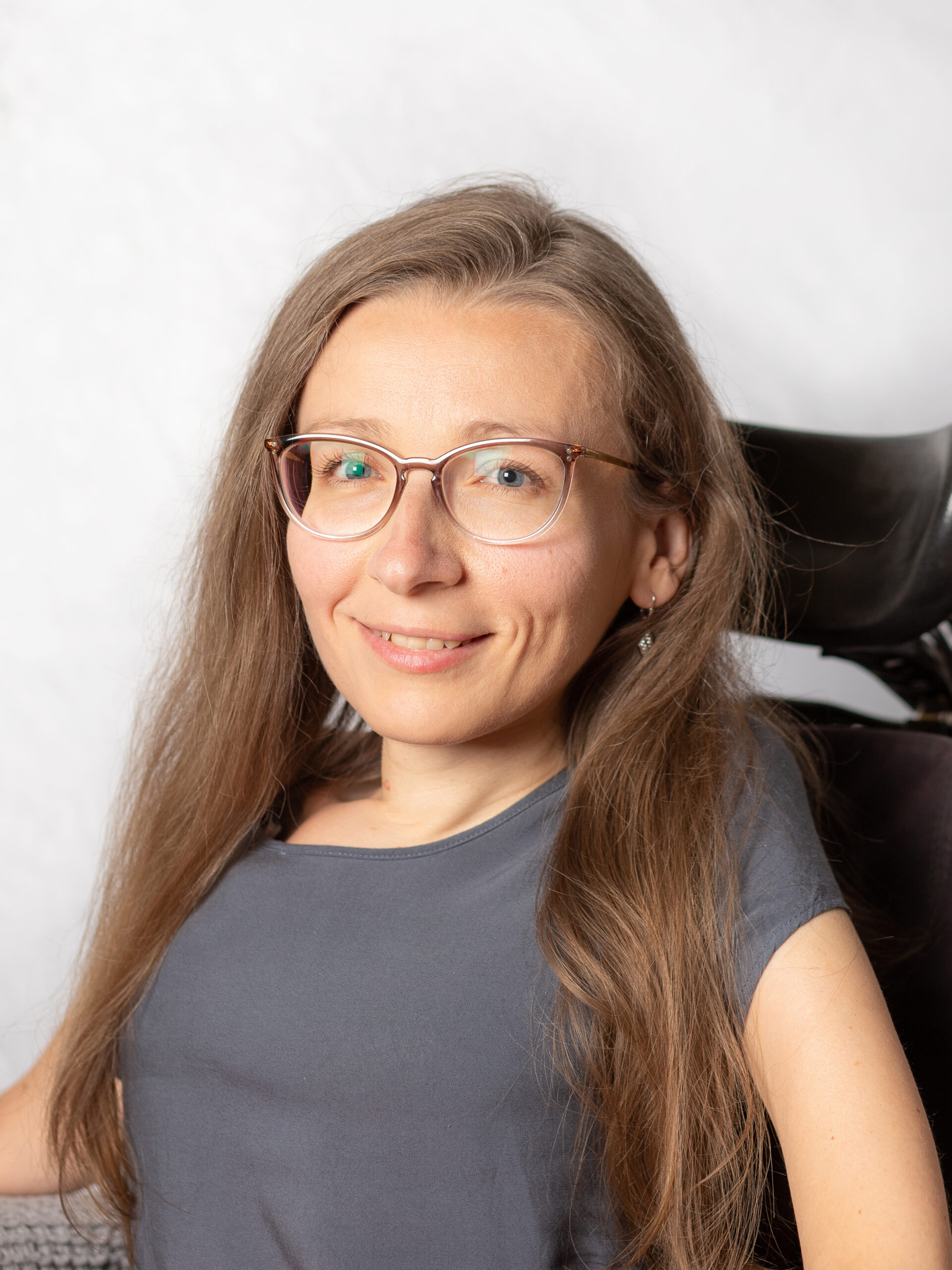 Frau mit langem, blondem Haar und Brille. Mit Lehne des Rollstuhls im Hintergrund.