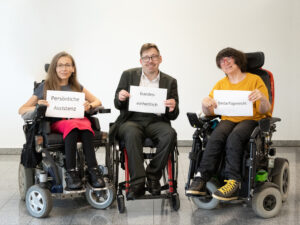 zwei Frauen und ein Mann im Rollstuhl halten jede:r ein Schild in der Hand. Darauf steht: Persönliche Assistenz - bundeseinheitlich - bedarfsgerecht