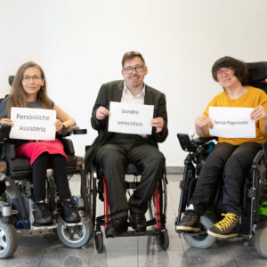 zwei Frauen und ein Mann im Rollstuhl halten jede:r ein Schild in der Hand. Darauf steht: Persönliche Assistenz - bundeseinheitlich - bedarfsgerecht