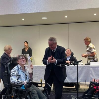 Roswitha Schachinger hält den Preis (eine Statue) in der Hand. Neben ihr steht ein Mann, der klatscht und gegenüber sitzt eine Rollstuhlfahrerin. Hinter ihr ein Podium mit mehreren Personen.
