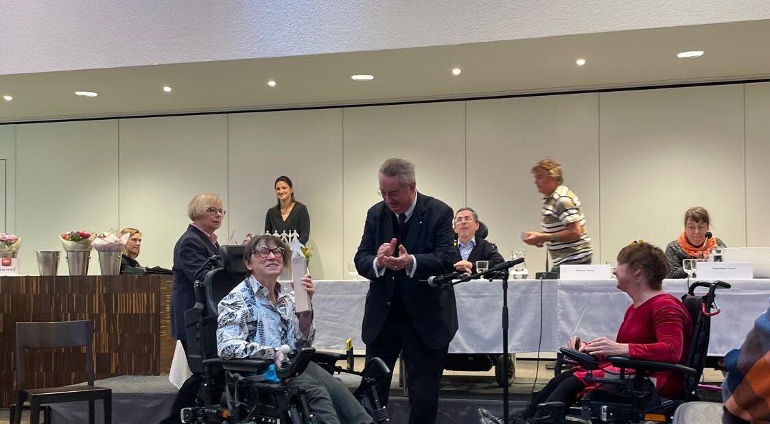 Roswitha Schachinger hält den Preis (eine Statue) in der Hand. Neben ihr steht ein Mann, der klatscht und gegenüber sitzt eine Rollstuhlfahrerin. Hinter ihr ein Podium mit mehreren Personen.