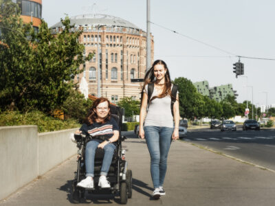Zwei junge Frauen auf der Straße vor den Gasometern. Eine sitzt im Rollstuhl.