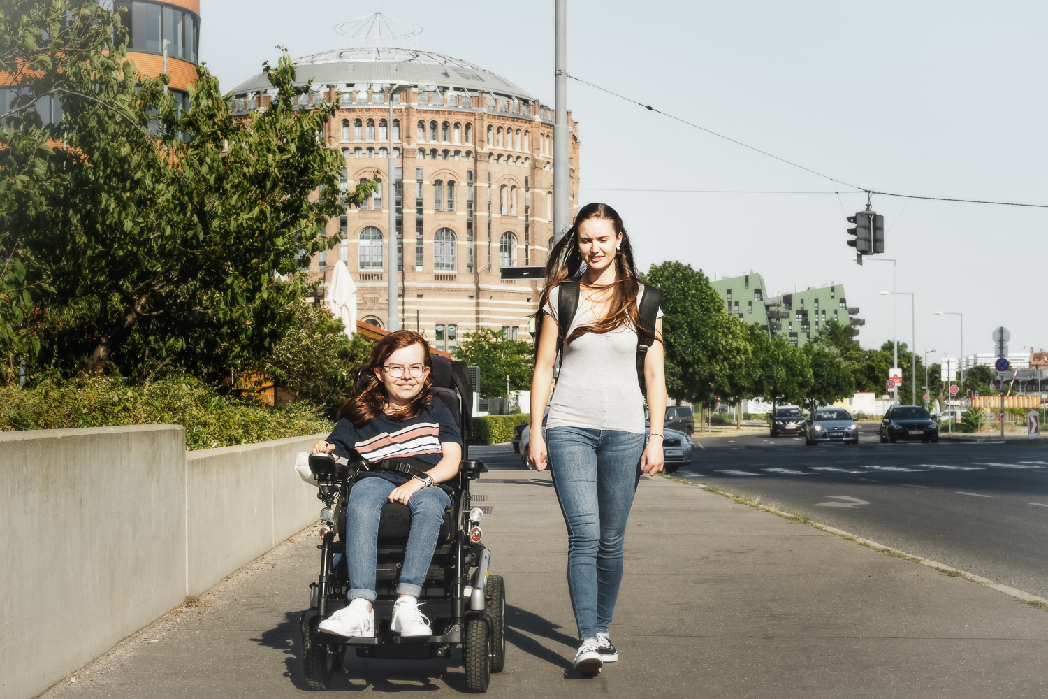 Zwei junge Frauen auf der Straße vor den Gasometern. Eine sitzt im Rollstuhl.
