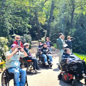 Mehrere Personen mit und ohne Rollstuhl im Wald. Einige halten ein Fernglas in der Hand. Sie beobachten etwas.
