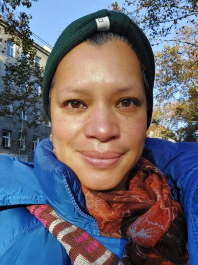 Frau mit Kopfbedeckung blickt in die Kamera. Neben ihr sind Bäume zu sehen.