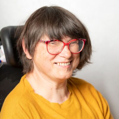 Frau mit halblangem, braunem Haar lächelt in die Kamera. Sie trägt eine rote Brille und einen gelben Pullover.