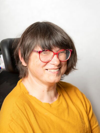 Frau mit halblangem, braunem Haar lächelt in die Kamera. Sie trägt eine rote Brille und einen gelben Pullover.