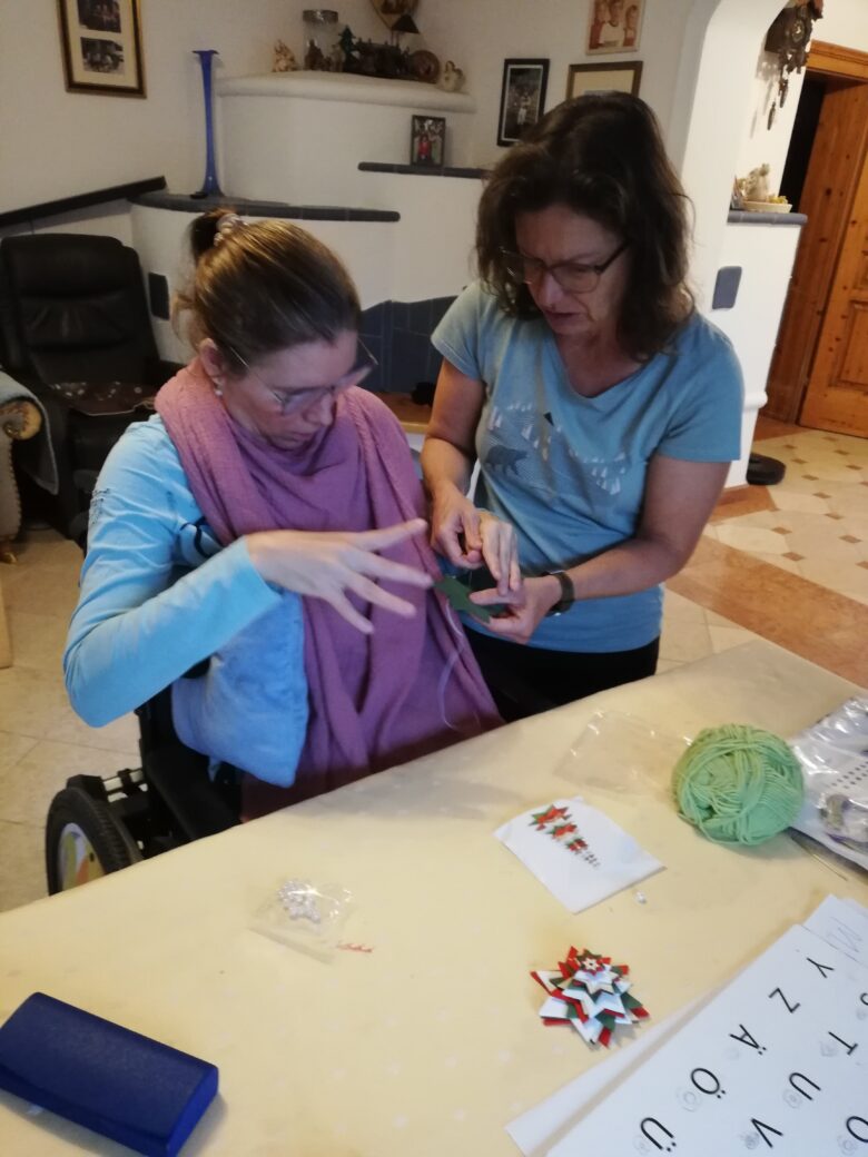 Zwei Frauen sitzen an einem Tisch und basteln. Eine Frau sitzt im Rollstuhl.