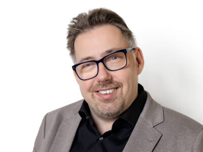 Mann trägt schwarze Brille und einen grauen Anzug. Er lächelt freundlich in die Kamera.