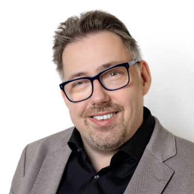 Mann trägt schwarze Brille und einen grauen Anzug. Er lächelt freundlich in die Kamera.