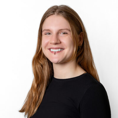 Frau mit brünetten, langen Haaren lächelt in die Kamera. Sie trägt ein Piercing.