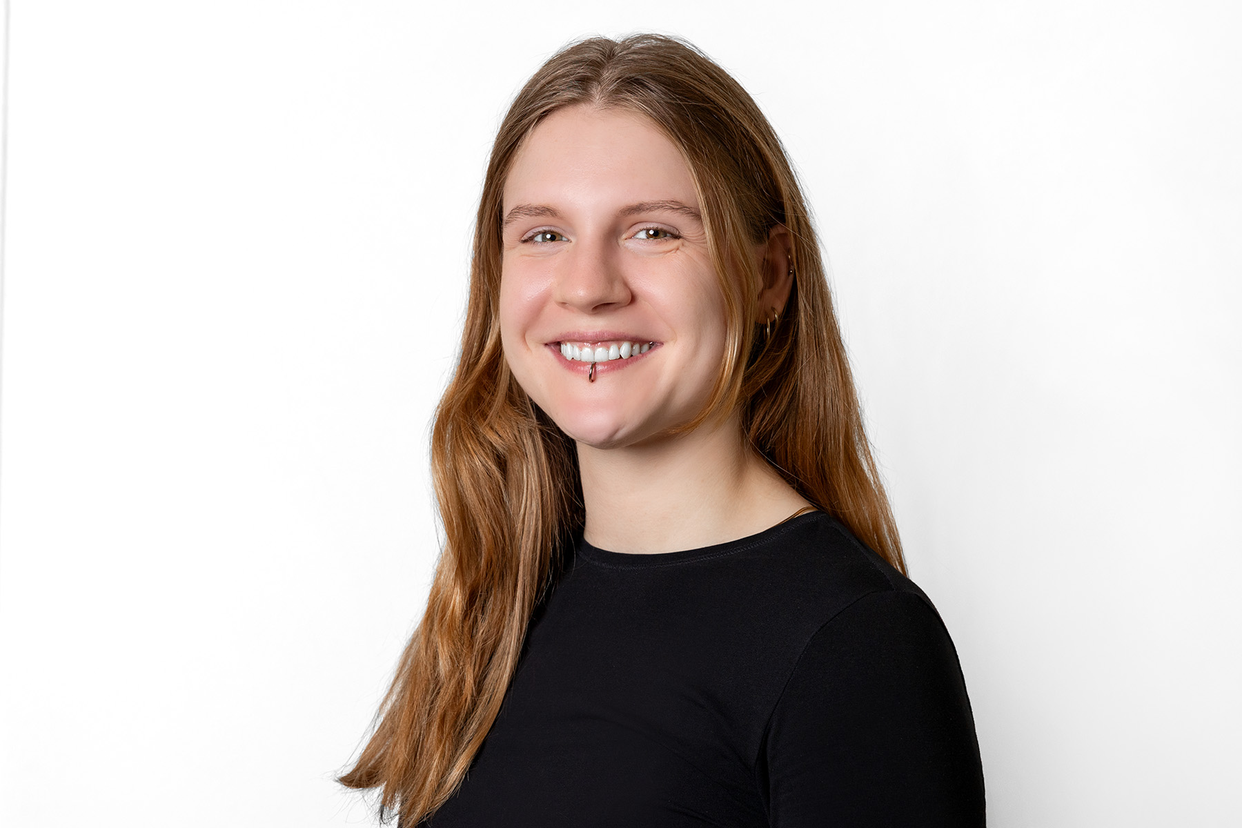 Frau mit brünetten, langen Haaren lächelt in die Kamera. Sie trägt ein Piercing.