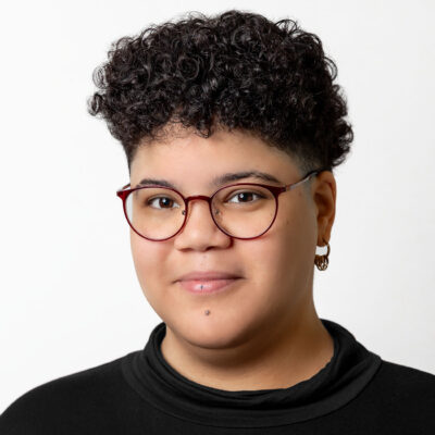 Person mit kurzem lockigen Haar und Brille lächelt in die Kamera