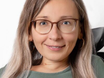 Frau mit langem Haar trägt eine Brille und lächelt freundlich in die Kamera