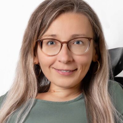 Frau mit langem Haar trägt eine Brille und lächelt freundlich in die Kamera