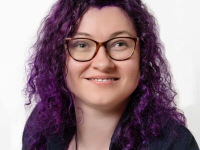 Frau mit dunklem, leicht violettem Haar. Sie trägt Brille.