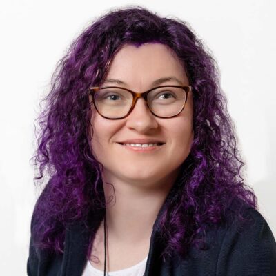 Frau mit dunklem, leicht violettem Haar. Sie trägt Brille.