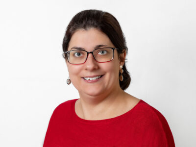 Frau mit schwarzem Haar in einem Zopf trägt ein rotes Oberteil und Brille. Sie lächelt in die Kamera