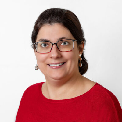 Frau mit schwarzem Haar in einem Zopf trägt ein rotes Oberteil und Brille. Sie lächelt in die Kamera