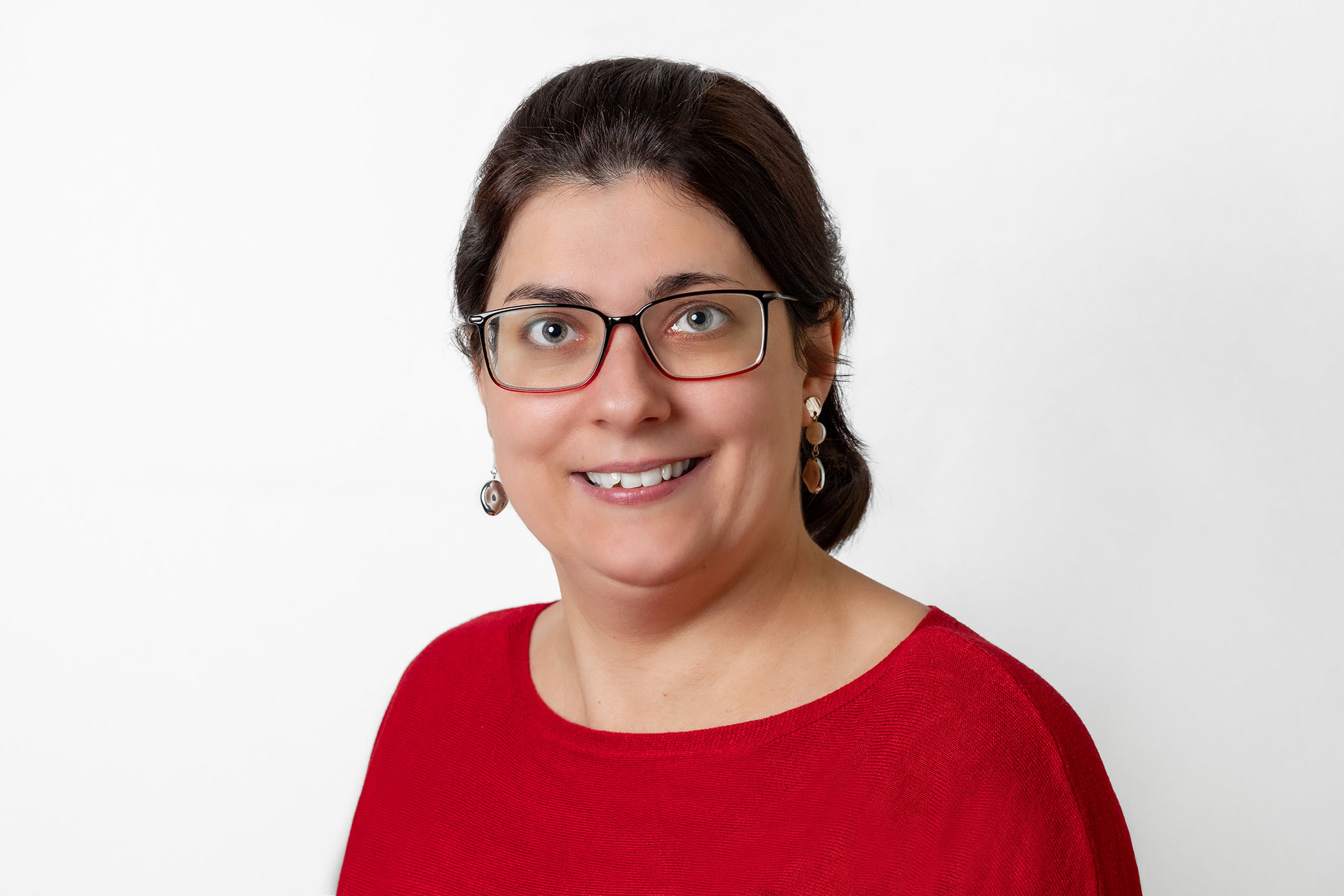 Frau mit schwarzem Haar in einem Zopf trägt ein rotes Oberteil und Brille. Sie lächelt in die Kamera