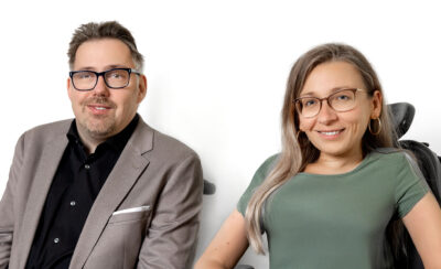 Eine Frau und ein Mann lächeln in die Kamera. Die Frau hat lange, blonde Haare, der Mann dunkles, kurzes Haar. Beide tragen eine Brille.